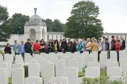 A group of people stands among rows of white headstones in a cemetery, with a domed memorial structure and trees in the background.
