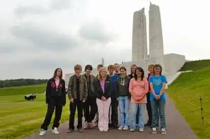 A group of people stands on a walkway in front of the Vimy Ridge Memorial, with green grass and cloudy skies in the background.