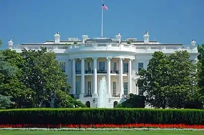 The White House in Washington, D.C., is seen from the front garden with a fountain, flowerbeds, and trees under a clear blue sky.