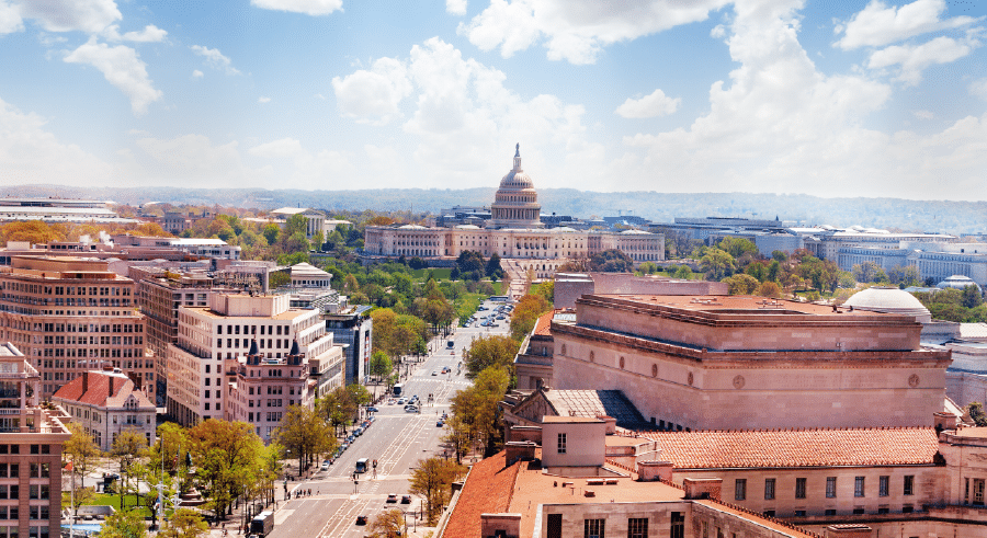 Aerial view of the United States Capitol building in Washington, D.C., with surrounding city buildings and tree-lined streets under a partly cloudy sky.