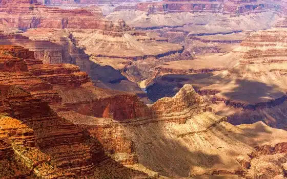 Aerial view of the Grand Canyon showing layered rock formations, steep cliffs, and deep valleys under sunlight and shadows.