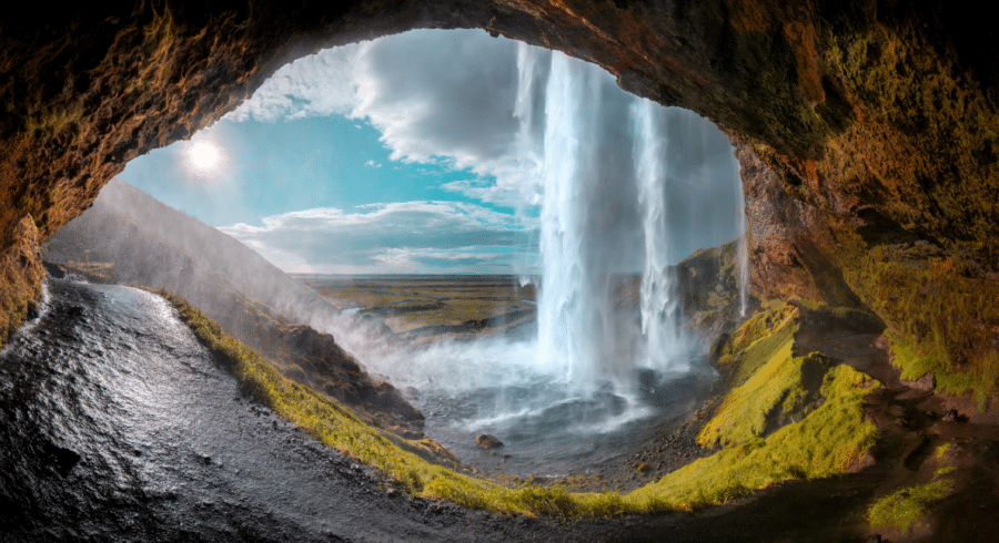 View from inside a cave behind a tall waterfall, with bright sunlight, mist, and green moss on the surrounding rocks and landscape.