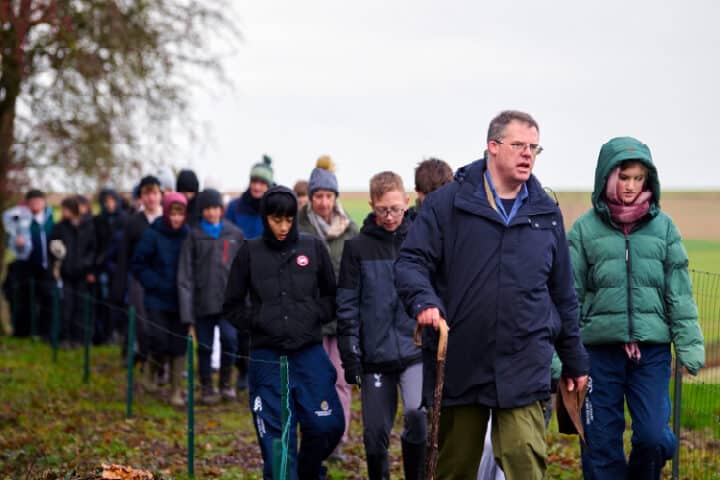 A group of people, bundled in winter clothing, walk in single file along a fenced path outdoors, led by a man holding a walking stick.