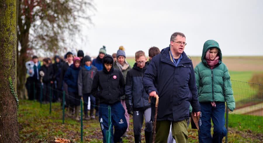 A group of people, led by a man with a walking stick, walk along a fenced dirt path in a rural area on a cloudy day.