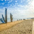 Metal sculptures stand on a sandy beach near the sea, with people walking nearby and flags flying beside stone steps under a partly cloudy sky.