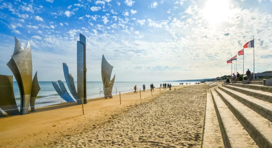 People walk along a sandy beach with metal sculptures, flagpoles with various flags, and stone steps under a bright sky.