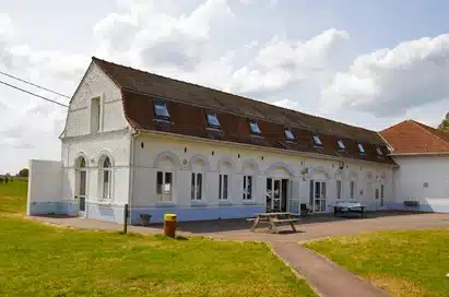 A white, two-storey building with arched windows and a red-tiled roof, featuring a table tennis table and benches outside on a sunny day.