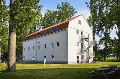 A three-storey white building with a red sloped roof, small windows, and an external metal staircase, surrounded by tall trees and grass.