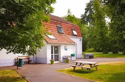 A white two-storey building with a brown sloped roof, several windows, a glass entrance door, and a bench outside, surrounded by grass and trees.