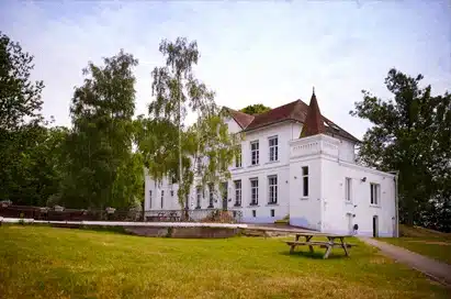 A large white two-storey building with multiple windows, surrounded by trees, a curved pathway, and decorative flower pots on a railing in the foreground.