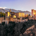 A large historic palace and fortress complex sits on a hill, surrounded by trees, with mountains visible in the background under a clear sky.