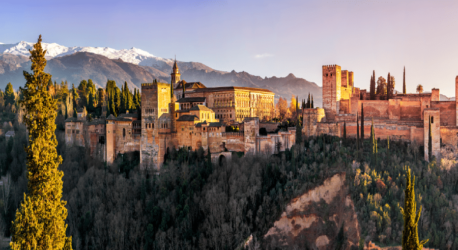 Panoramic view of the Alhambra palace and fortress complex in Granada, Spain, with mountains and trees in the background under a clear sky.