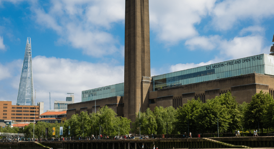 The Tate Modern art museum stands by the River Thames in London, with the Shard skyscraper visible in the background under a partly cloudy sky.