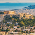 View of the Acropolis with the Parthenon on a hill in Athens, Greece, surrounded by city buildings and greenery, with the sea visible in the background.