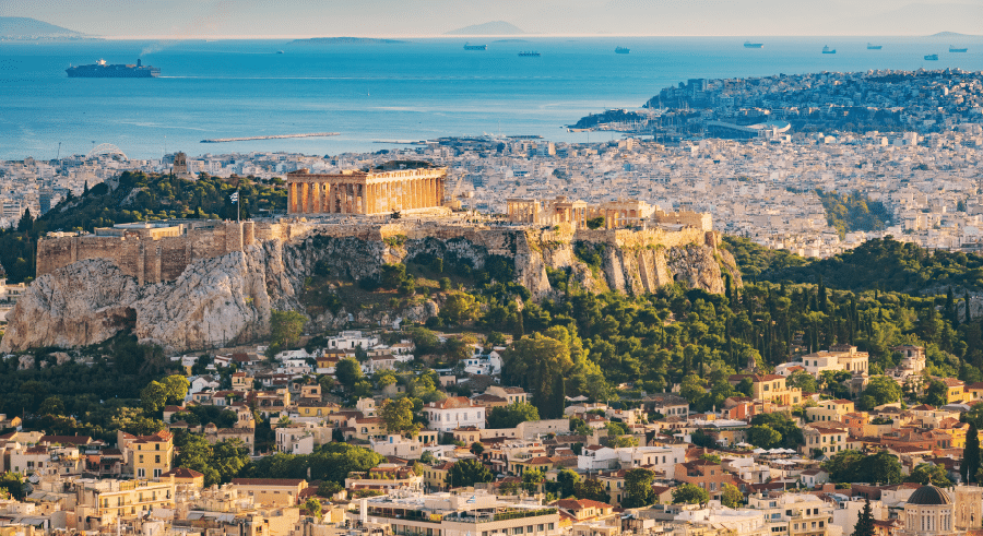 The Acropolis stands on a rocky hill above the city of Athens, surrounded by urban buildings, with the sea and distant ships in the background.