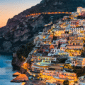 Coastal hillside town with colourful buildings illuminated at dusk, overlooking calm sea and steep cliffs.