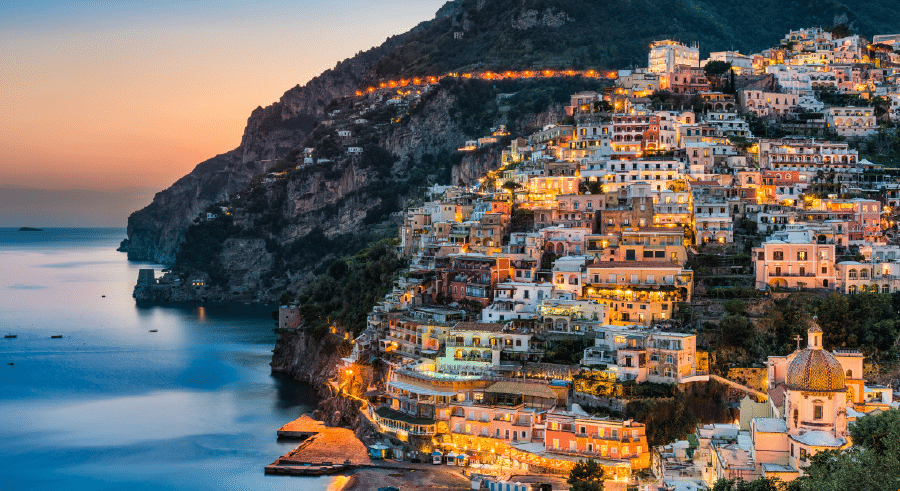 A coastal hillside town with colourful buildings, illuminated at dusk, overlooking calm blue water and steep, forested cliffs.