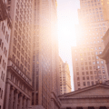 Sunlight shines between tall office buildings in a city financial district, with American flags visible on some of the buildings.