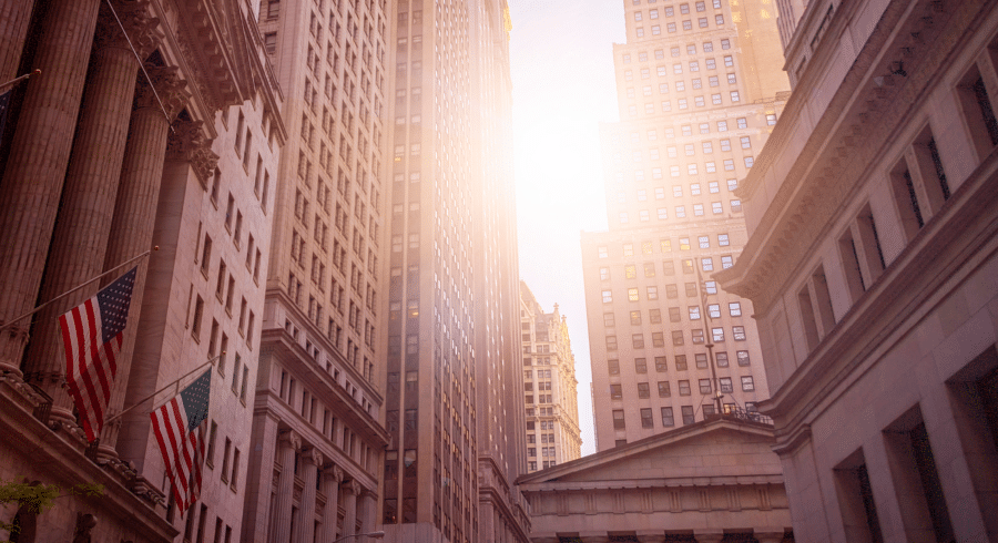 Tall office buildings line a narrow street with American flags displayed, as sunlight shines between the skyscrapers in an urban cityscape.