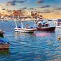 Small boats float on a calm harbour with a distant coastal town and partly cloudy sky at sunset.