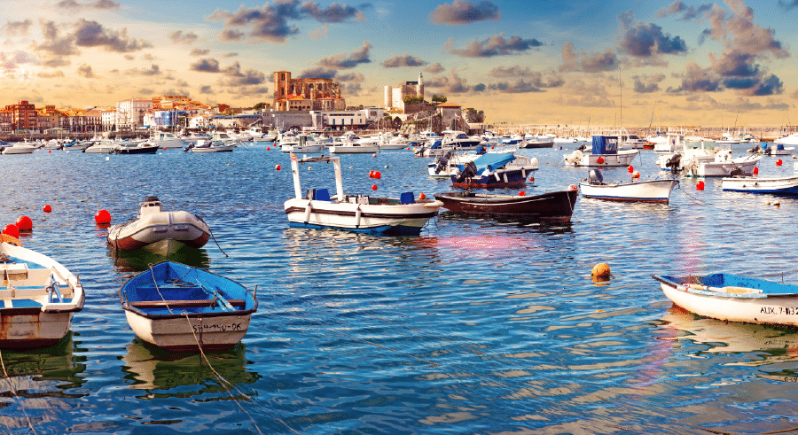Small boats floating on calm harbour waters with a coastal town and historic buildings in the background under a partly cloudy sky at sunset.