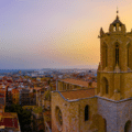 Aerial view of a historic stone church tower overlooking a cityscape at sunset, with the sea visible in the distance.