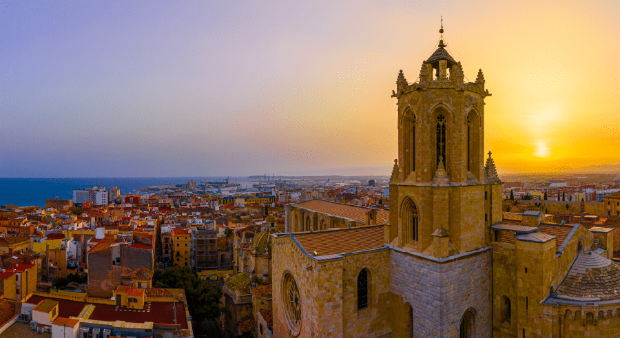 Panoramic view of a historic stone cathedral tower overlooking a cityscape at sunset, with the sea in the background.