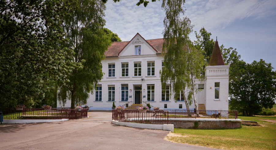 A large white two-storey house with a pointed turret, surrounded by trees and a curved drive under a partly cloudy sky.