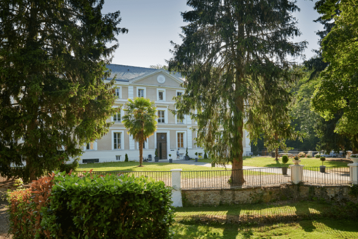 A large white mansion with columns stands behind a fenced garden, surrounded by tall trees and greenery on a sunny day.