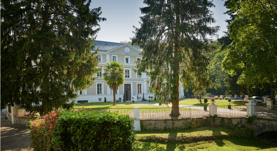 A large white mansion with columns stands behind a fenced garden, surrounded by tall trees and greenery on a sunny day.