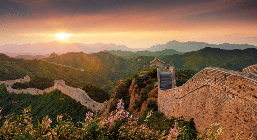 The Great Wall of China stretches across green hills under a colourful sunset sky, with wildflowers in the foreground.