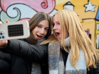 Two young people in winter coats and scarves take a selfie together in front of a colourful mural, both smiling with surprised expressions.