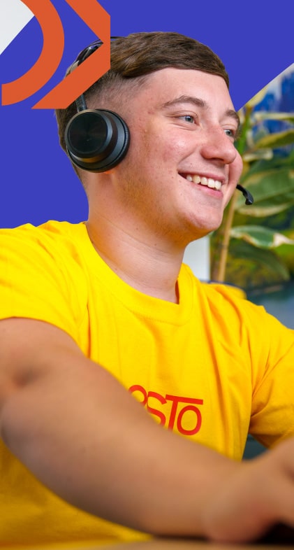 A young person wearing a yellow shirt and headset smiles while looking at a computer screen, with a plant and blue background behind them.