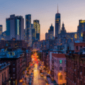City skyline at dusk with tall modern skyscrapers in the background and a street lined with older brick buildings and cars in the foreground.