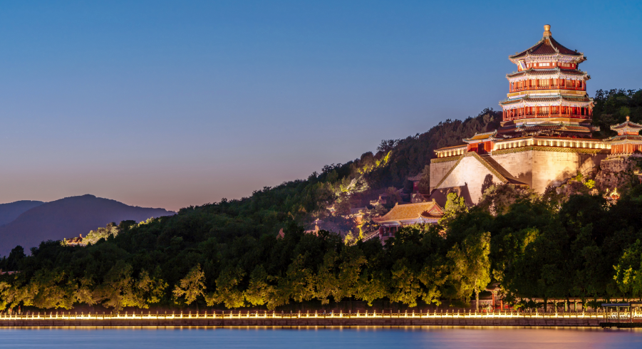 A traditional Chinese pavilion on a hillside is illuminated at dusk, overlooking a line of trees and a calm body of water.