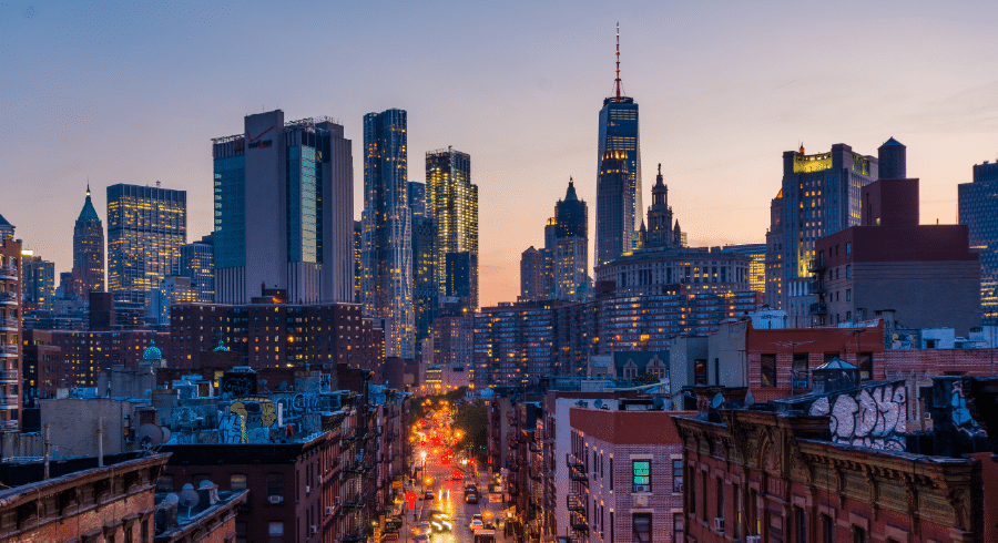 City skyline at dusk with tall buildings and lights, viewed from above a busy street with cars and graffiti on nearby rooftops.