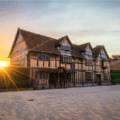 Historic half-timbered building with a steep roof at sunset, light shining over the hedge, on a paved street with clear sky.