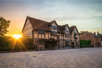 Historic half-timbered building with a steep roof at sunset, light shining over the hedge, on a paved street with clear sky.