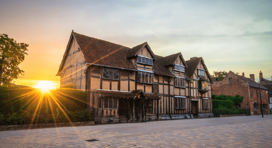 A timber-framed historic building with leaded windows and a steep roof, pictured at sunset with sunlight shining from the left.