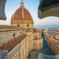 View of Florence Cathedral’s red-tiled dome and surrounding cityscape, seen through a decorative stone opening, under a clear sky.