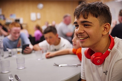 Teen boy wearing red headphones and a white T-shirt sits at a table with others in a casual indoor setting, smiling.