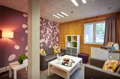A modern waiting room with grey armchairs, a white coffee table, a white cubed shelf with documents, and purple-accented walls and decor.