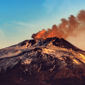 A volcano with snow-covered slopes releases a plume of dark smoke into the sky at sunrise or sunset.