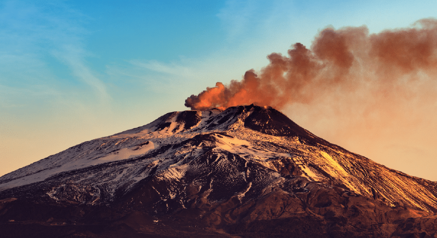 A volcano with snow on its slopes emits a thick plume of dark smoke into a blue and orange sky.