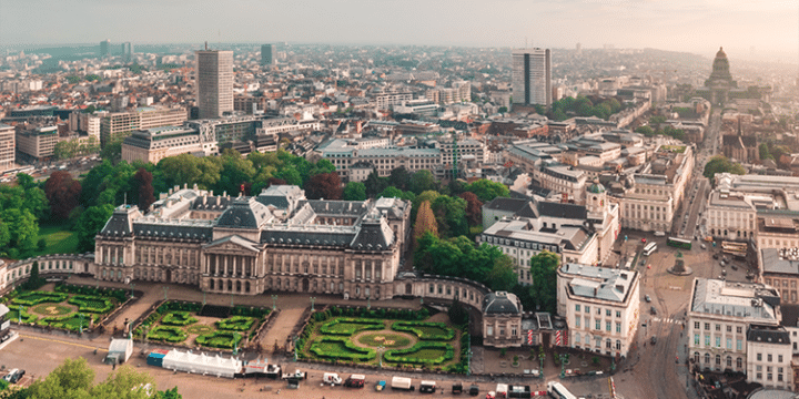 Aerial view of a large historic palace with manicured gardens in a city surrounded by modern and older buildings under a hazy sky.