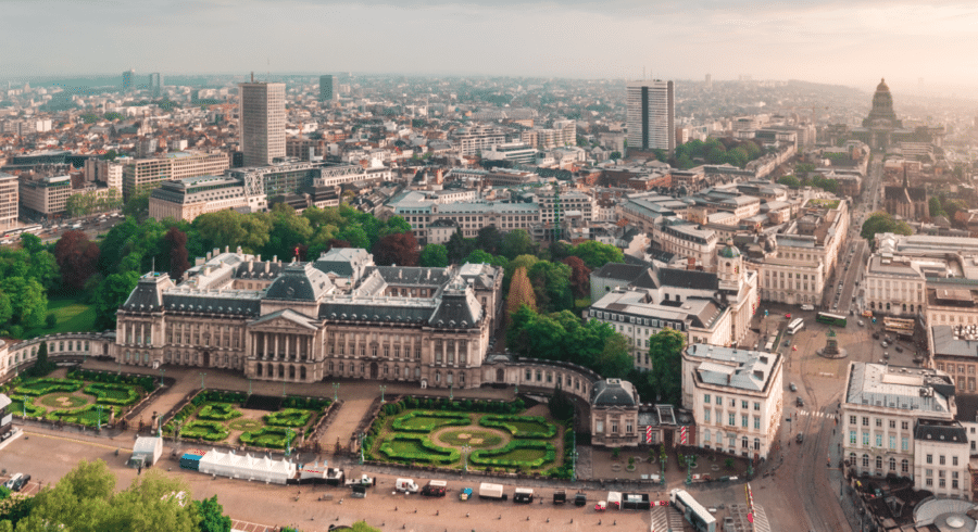 Aerial view of the Royal Palace of Brussels surrounded by formal gardens, with city buildings and streets extending into the distance.