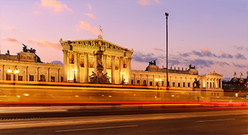 A neoclassical building with statues on the roof, illuminated at twilight, with blurred motion of traffic in the foreground.