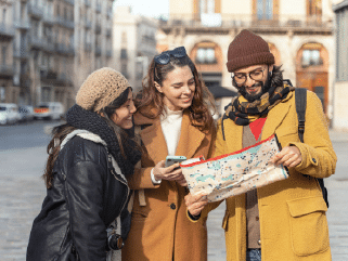 Three people in winter clothing stand outdoors in a city, looking at a map and a mobile phone, appearing to navigate or plan their route.