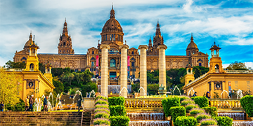 The image shows the National Palace in Barcelona, Spain, with cascading fountains, four columns, and a clear blue sky.