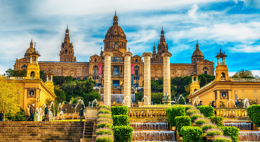 The National Art Museum of Catalonia in Barcelona, Spain, with four columns, fountains, and symmetrical gardens in front under a partly cloudy sky.